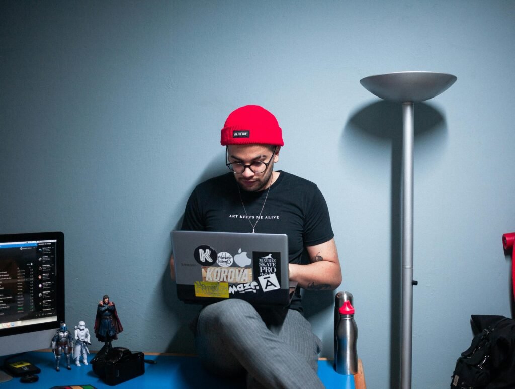 Freelancer in a red beanie working on a laptop indoors with decorative action figures and tech setup.