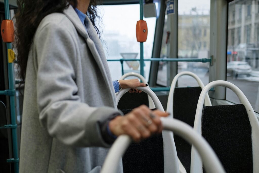A person holding a rail inside a city bus, wearing a gray coat, with a street view outside.